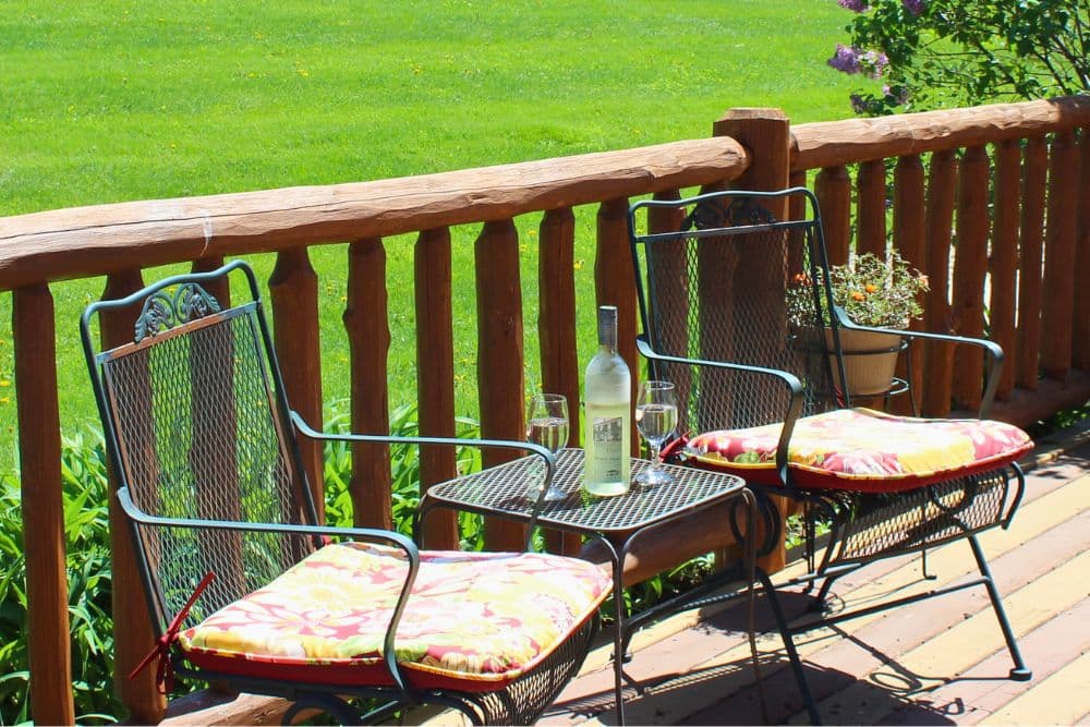 A sunny porch features two chairs with colorful cushions and a table holding a bottle of wine and glasses.