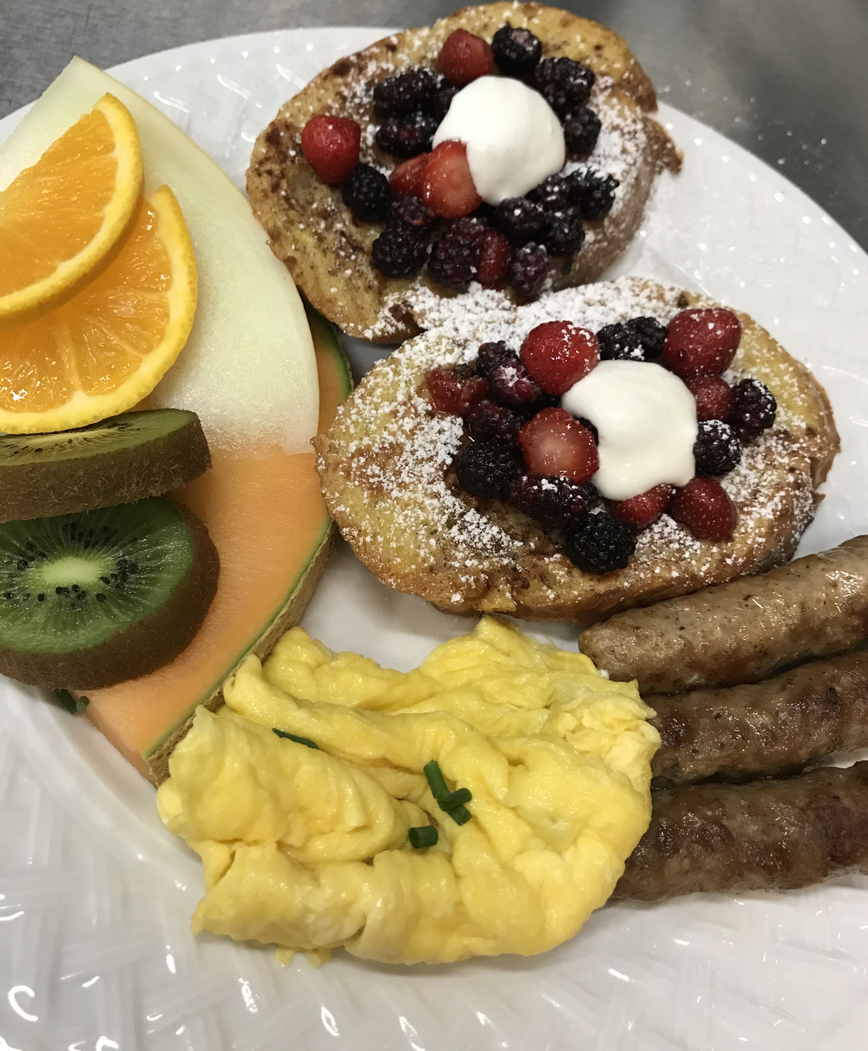 A plated breakfast featuring French toast topped with mixed berries and cream, scrambled eggs, sausage links, and assorted fresh fruit.
