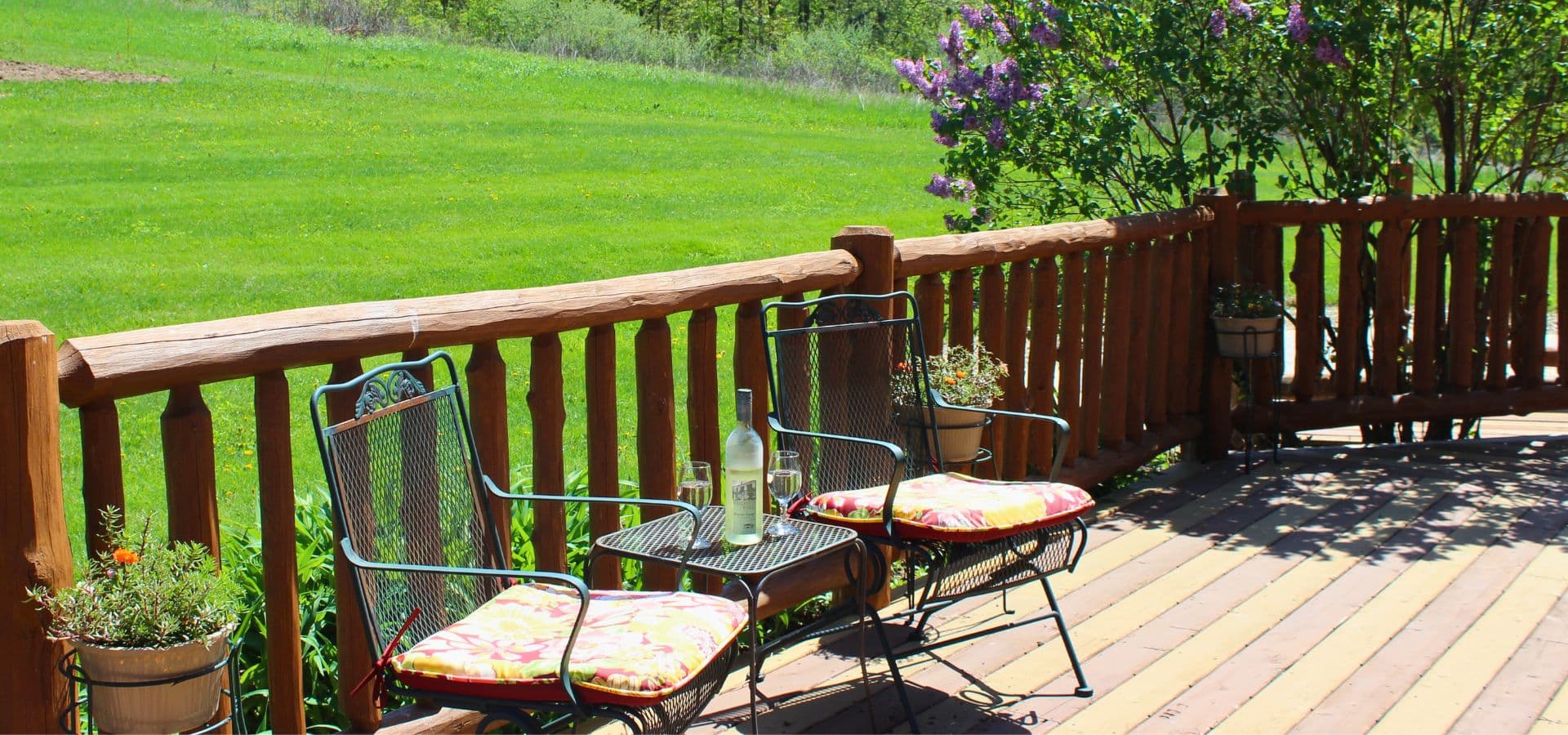 Two patio chairs with colorful cushions and a bottle of wine sit on a wooden deck overlooking a green field.